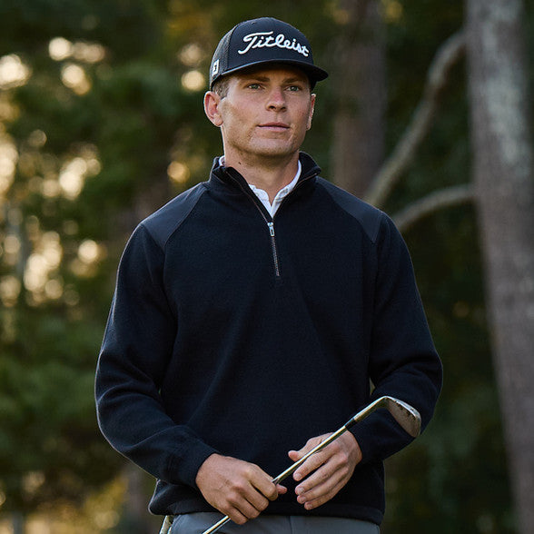 Man wearing a black Titleist cap and dark jacket holding a golf club with a blurred natural background